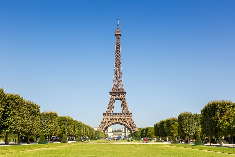 Ang Eiffel Tower, isang mataas na toreng gawa sa bakal na sala-sala, na makikita sa kahabaan ng damuhan na may mga puno sa ilalim ng malinaw na asul na kalangitan
