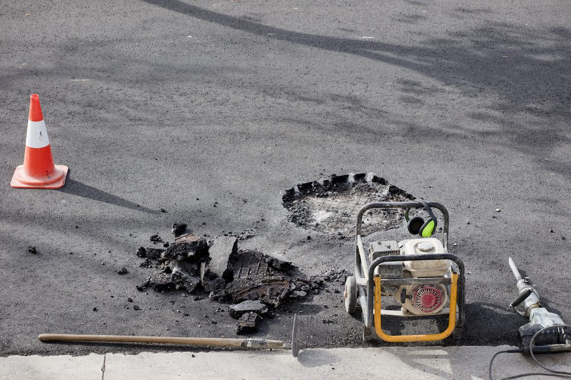 Sebuah lubang yang dibuat di jalan dengan satu kerucut lalu lintas, aspal yang pecah, sebuah generator, dan sebuah alat pemecah beton yang tertinggal di jalan.