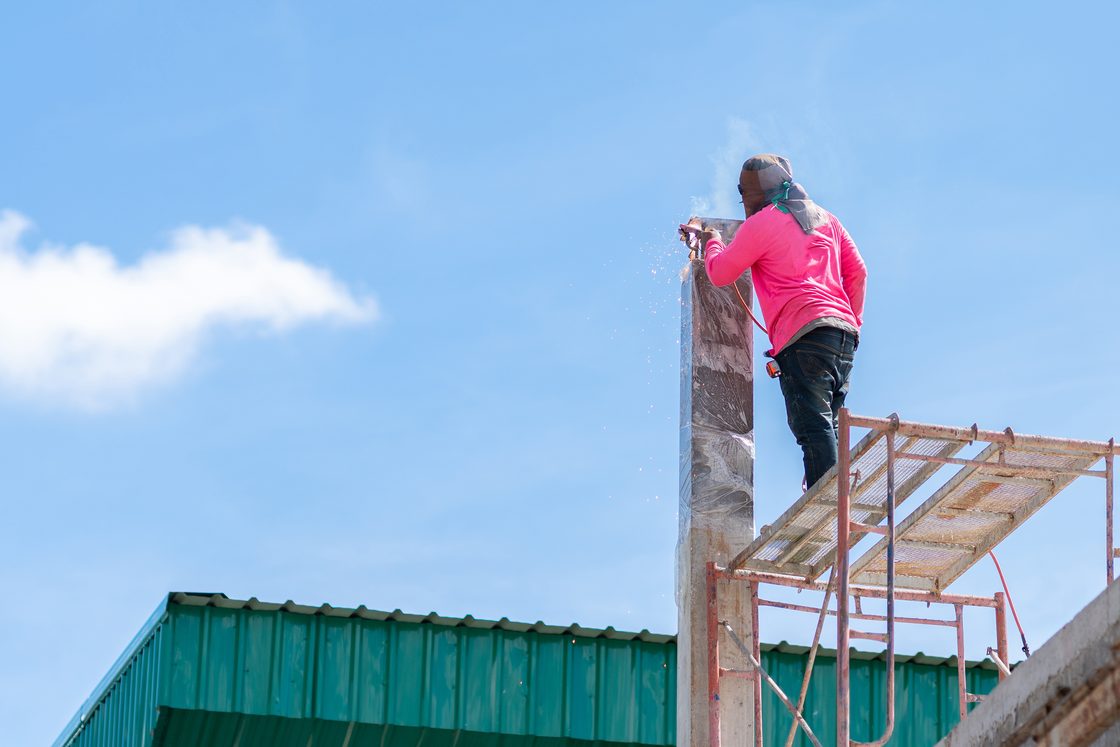 Seorang pekerja berdiri di atas platform perancah tanpa pagar pengaman, memotong bagian atas kolom beton sementara percikan api beterbangan.