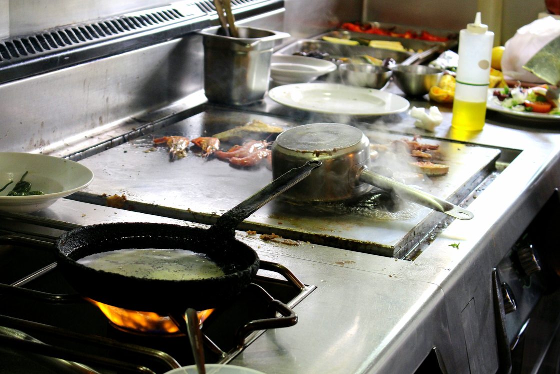 Sebuah kompor restoran dengan wajan gosong di atas api gas, panci di atas panggangan, dan botol minyak di dekatnya.