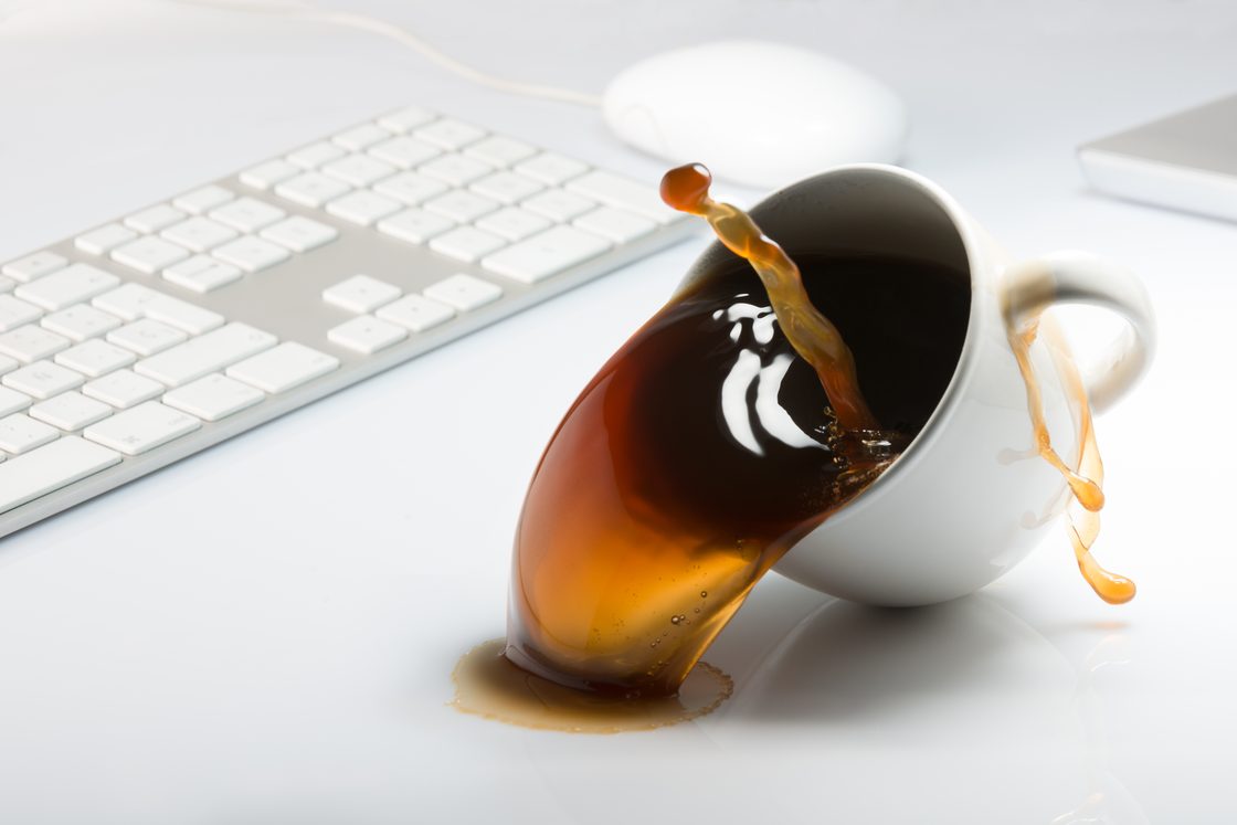 Secangkir kopi tumpah di samping keyboard dan mouse.
