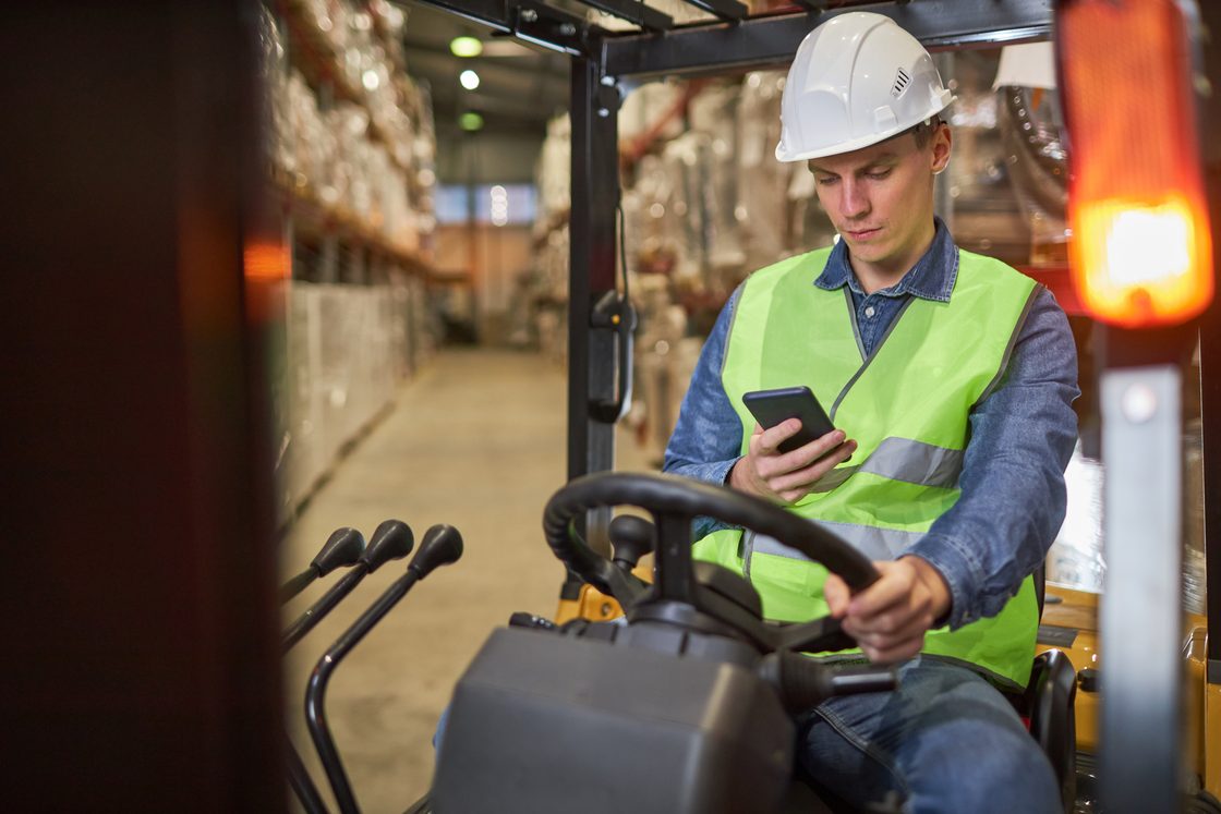 Seorang pengemudi forklift mengenakan helm pengaman sedang membaca ponselnya dengan satu tangan di kemudi.