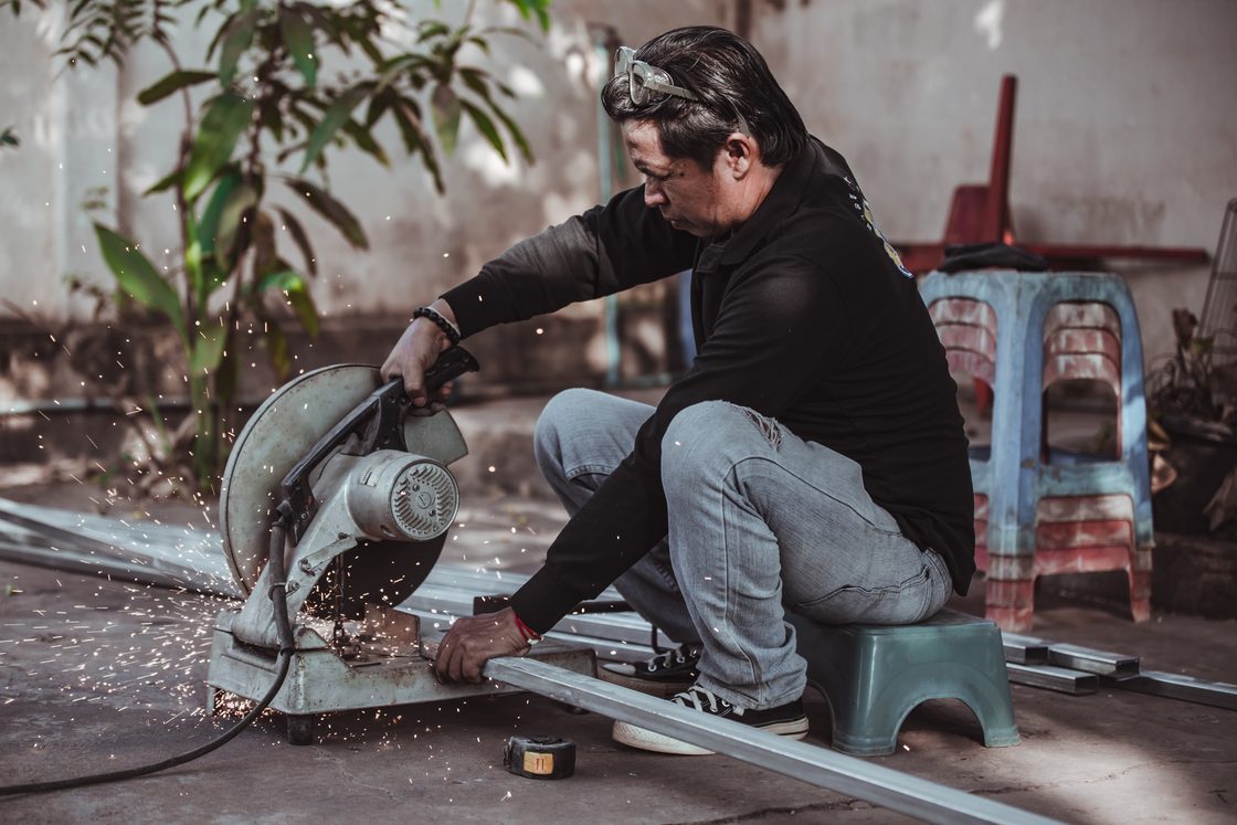 Seorang pria berjongkok di atas bangku rendah sambil memotong pipa baja dengan gergaji potong, percikan api beterbangan dan kacamata pengaman terdorong ke atas kepalanya.