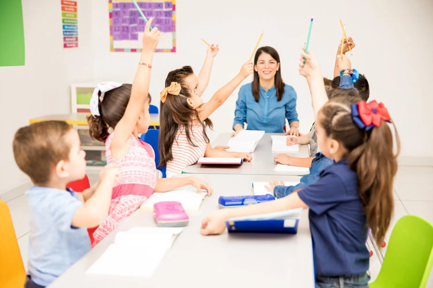 A nursery staff watching the children raise their hands in a classroom