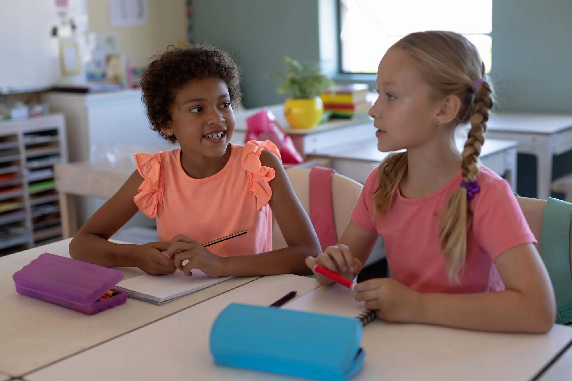 To unge skolepiger sidder sammen ved et klasseværelsesbord, taler og deler ideer under en time