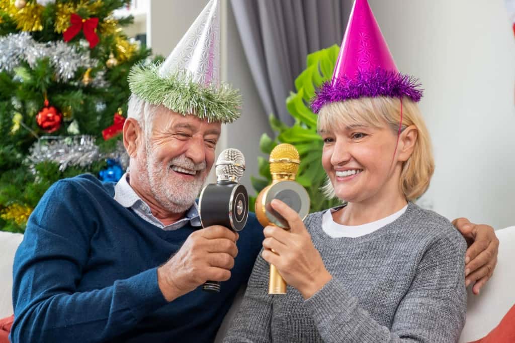 Older couple in party hats singing Christmas songs into karaoke microphones by the tree