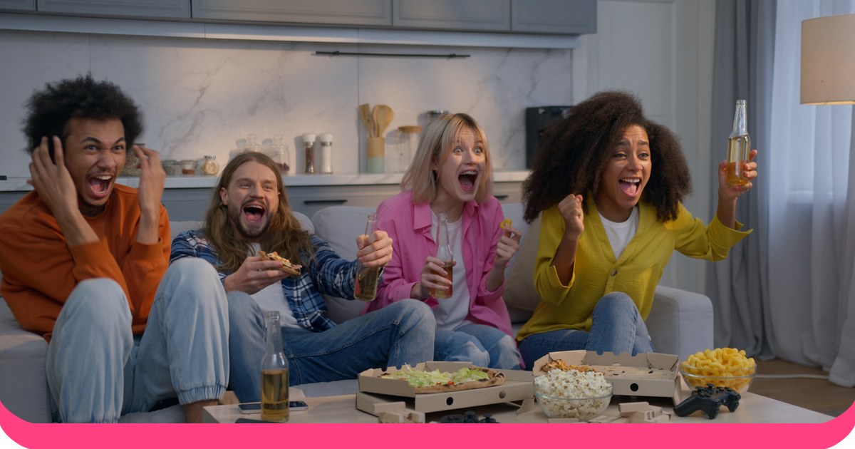 A group of friends cheering and laughing together on a sofa, with pizza and snacks on the table