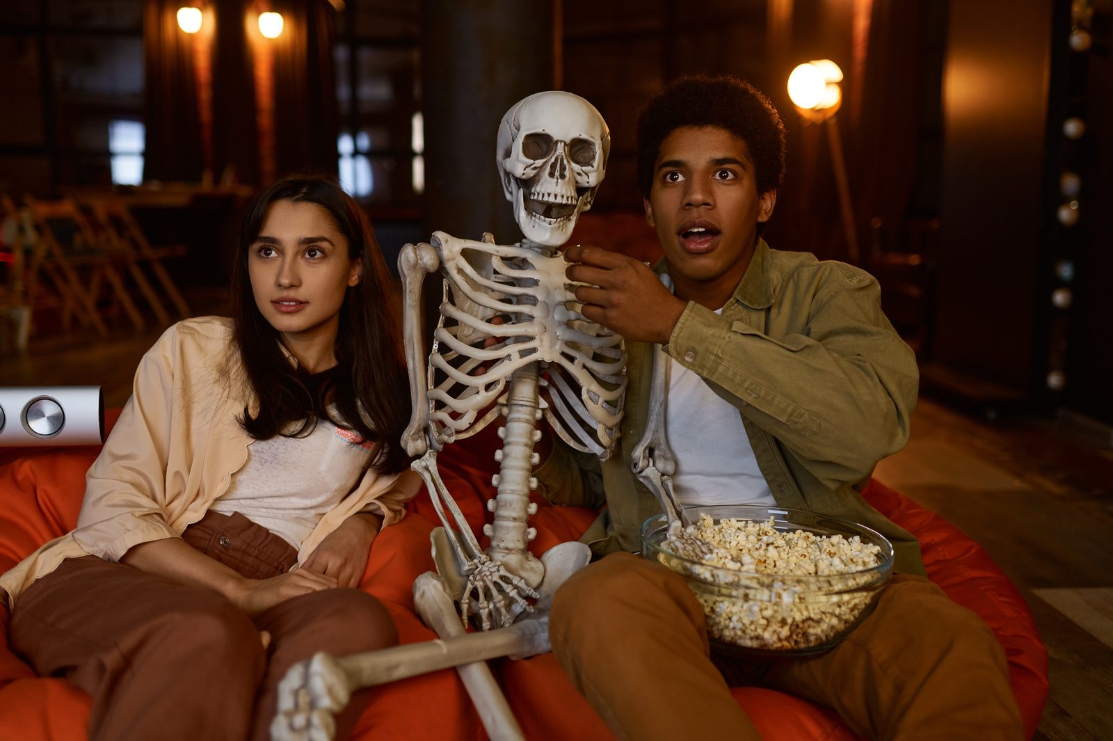 Zwei Freunde sitzen auf Sitzsäcken und schauen sich einen Horrorfilm an. Zwischen ihnen steht eine Schüssel Popcorn und ein Plastikskelett.
