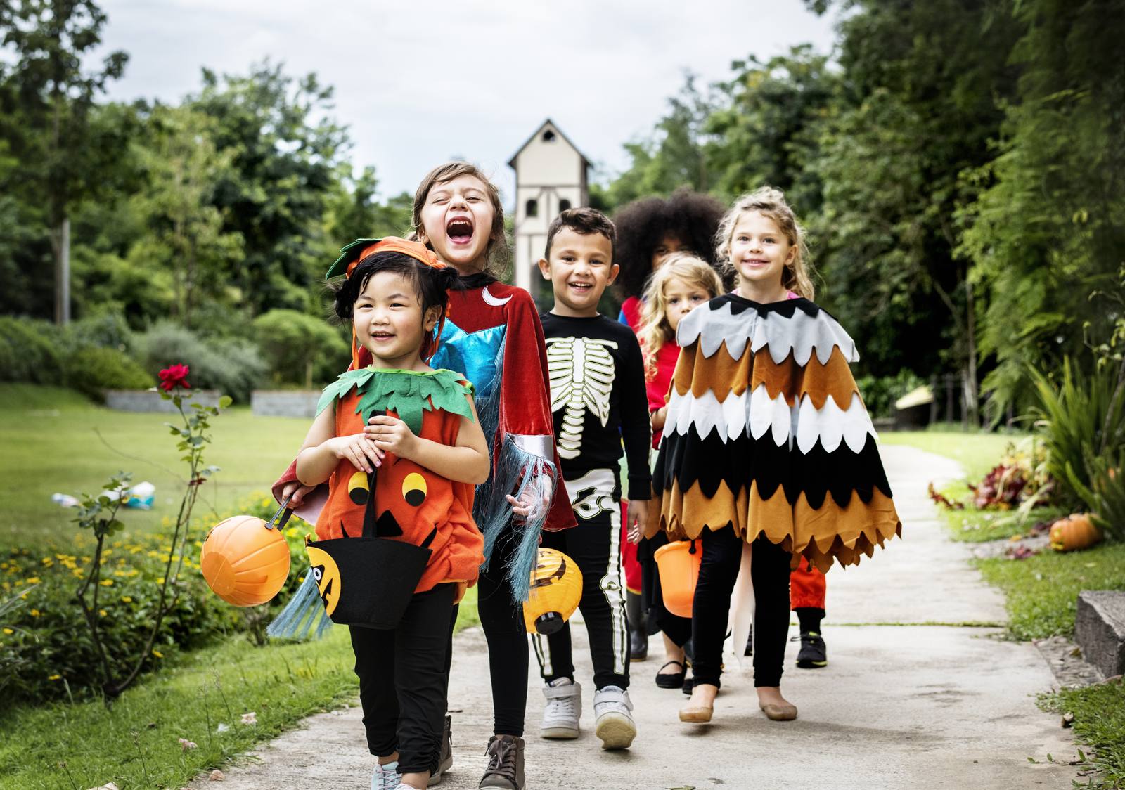 Eine Gruppe kleiner Kinder in Kürbis-, Skelett- und Vogelkostümen läuft mit Eimern voller Süßigkeiten einen Gartenweg entlang.