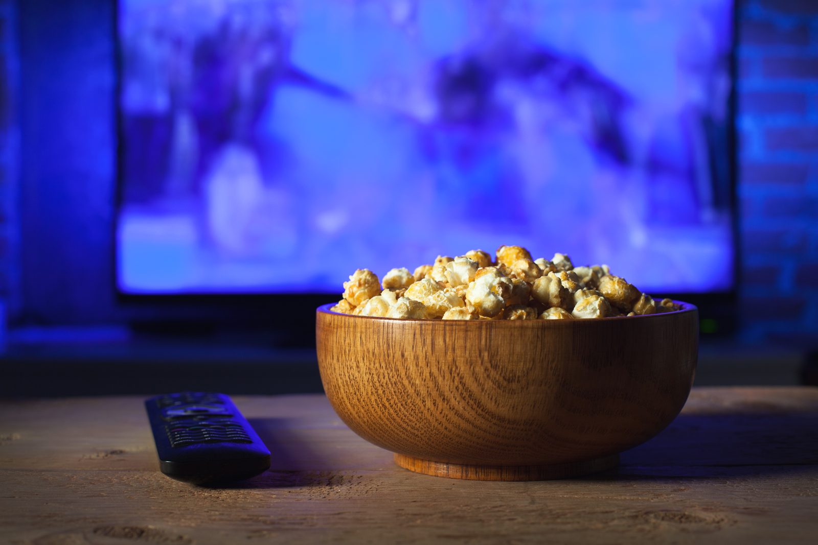 光るテレビの前のテーブルの上に、キャラメルポップコーンの入った木製のボウルとテレビのリモコンが置かれている。