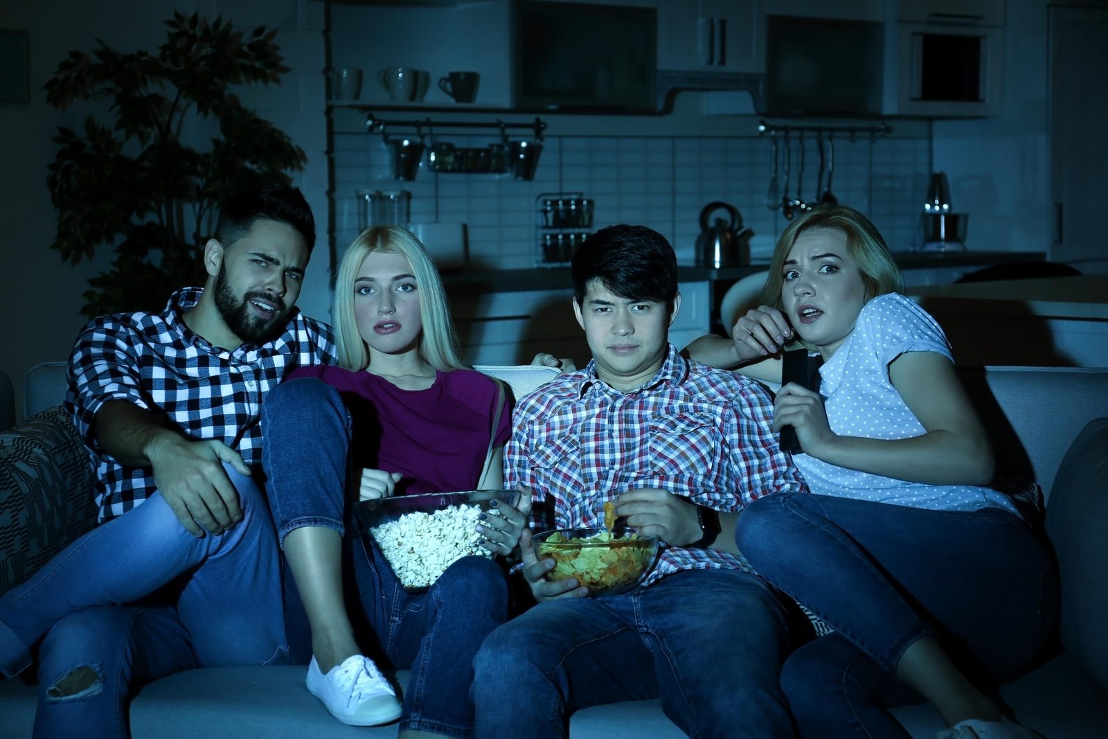 暗いリビングルームのソファに座った4人の友人が、ポップコーンやスナック菓子を食べながら、緊張した面持ちでホラー映画を観ている。