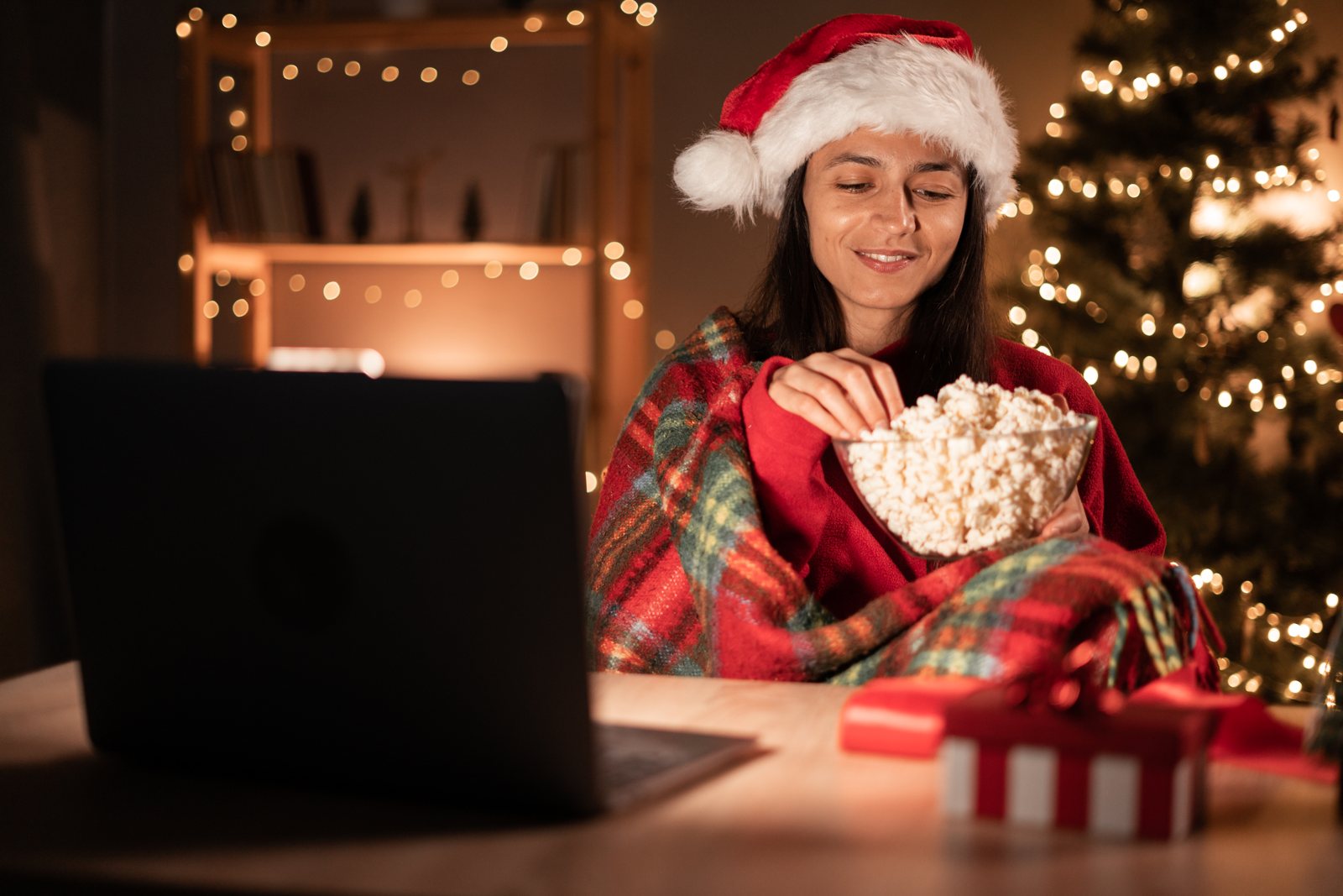 Woman in a Santa hat sitting under a blanket with a bowl of popcorn in front of a laptop, with Christmas lights behind her