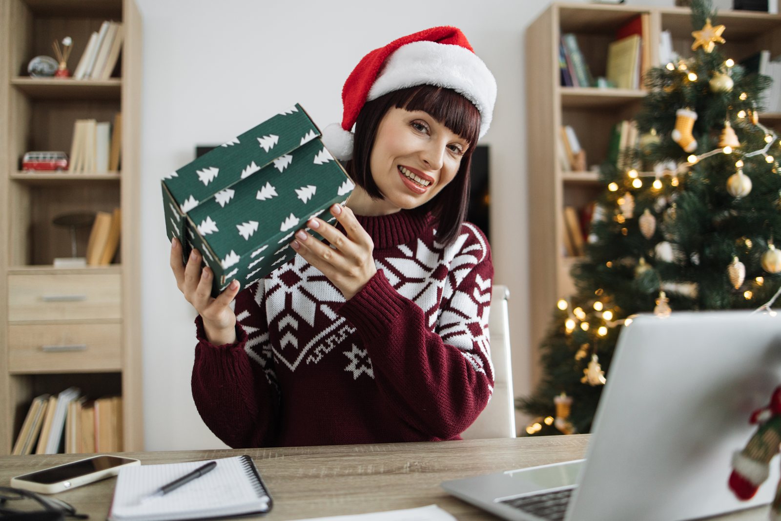 Woman in a Santa hat opening a gift box at her desk with a laptop open beside a decorated Christmas tree