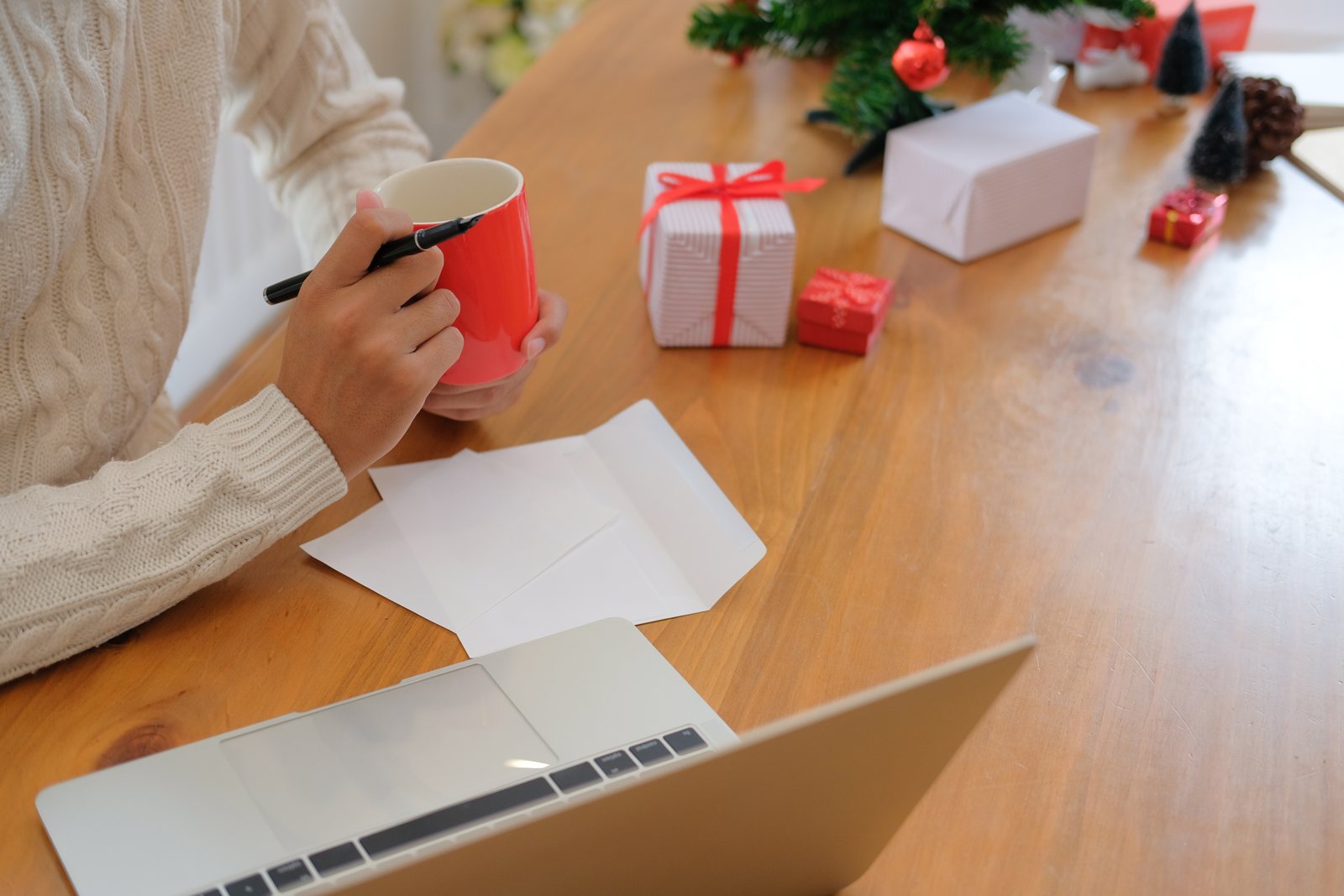 Person writing a Christmas card at a wooden table with a red mug, a laptop, wrapped presents and a small tree