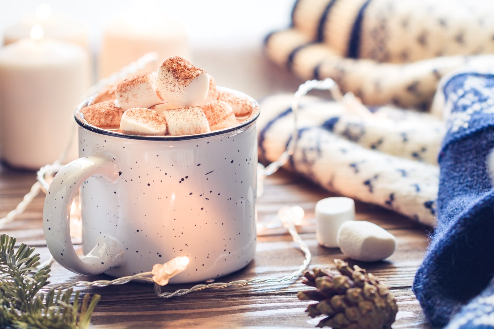 White enamel mug of hot chocolate topped with marshmallows beside candles, knitted socks and pine cones