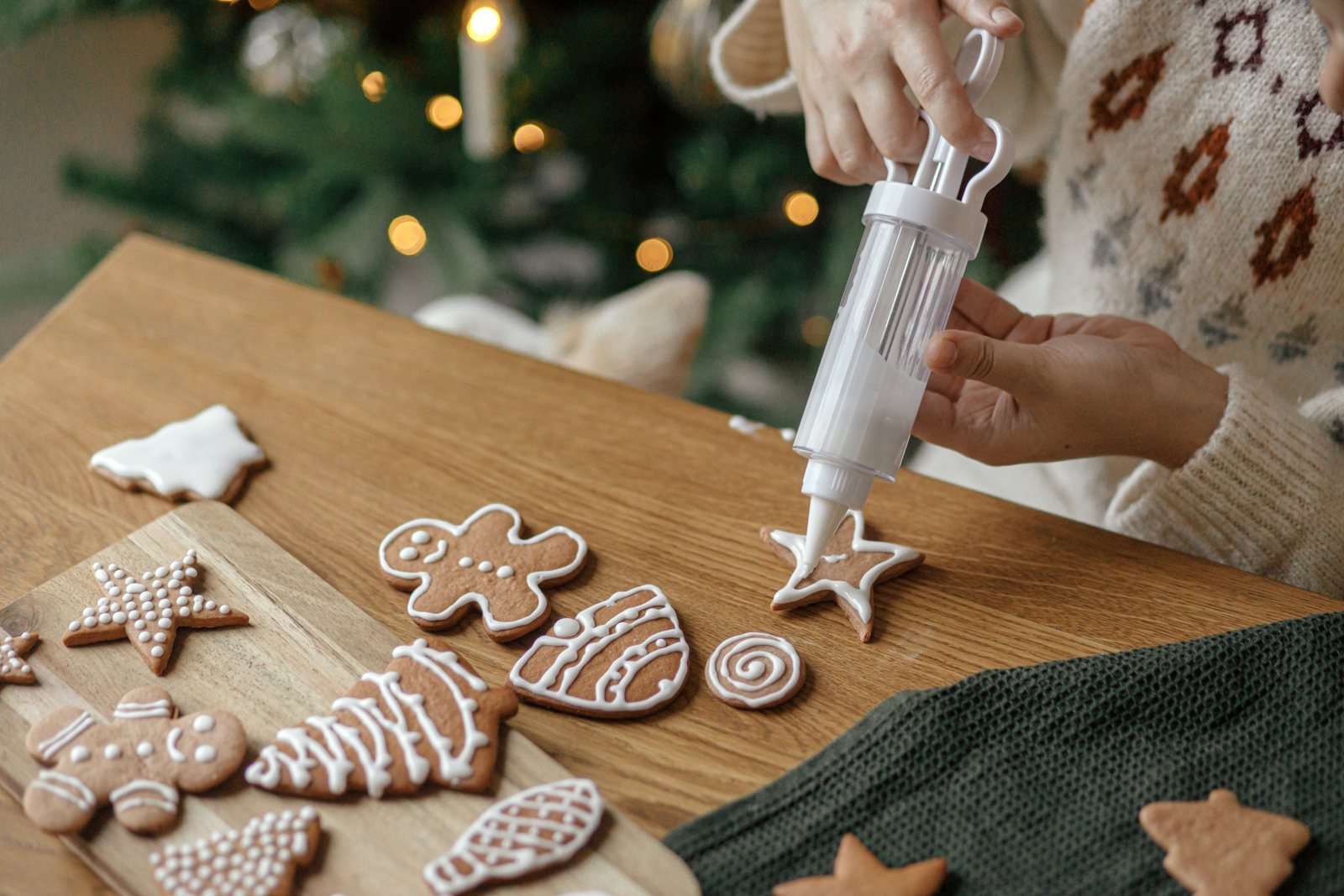 Hands piping white icing onto gingerbread biscuits on a wooden table beside a decorated Christmas tree