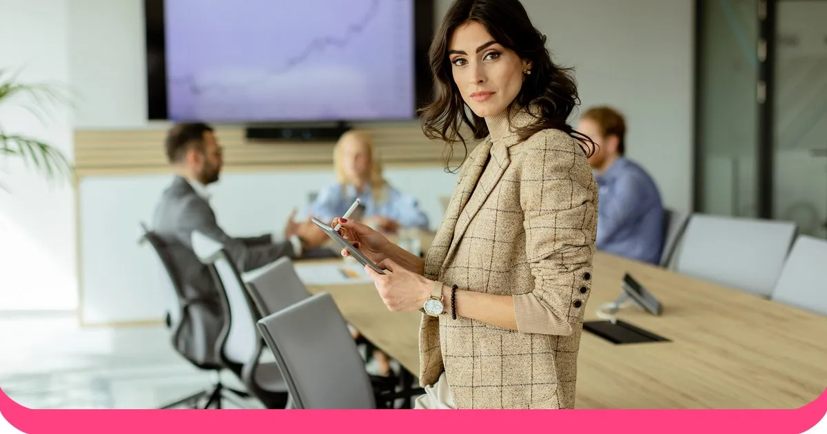 Businesswoman in a tweed blazer holding a tablet in a meeting room with colleagues behind her