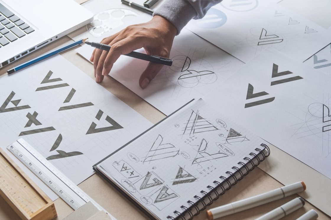 A designer's hand sketching rough black-and-white logo shapes in a notebook and on loose sheets