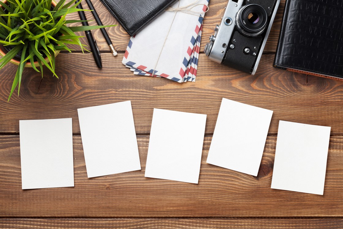 Five blank instant photo prints in a row on a wooden desk beside a vintage camera and a bundle of airmail envelopes