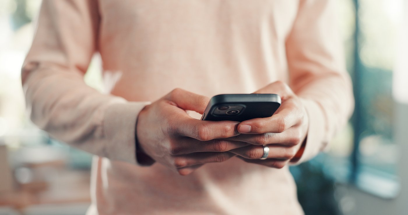 Pembe kazak giymiş, iki eliyle telefon tutan ve mesaj göndermek üzere olan bir kişinin yakın çekim görüntüsü.