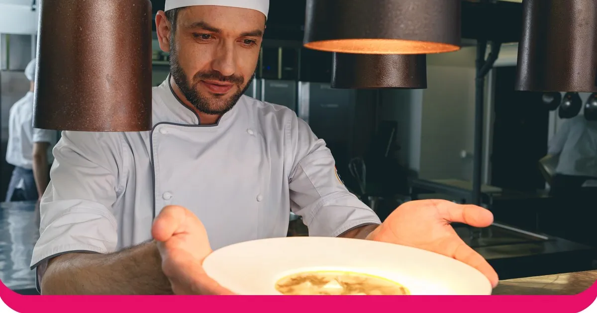 Ein Koch in weißer Kochkleidung hält in einer Restaurantküche ein angerichtetes Gericht zur Begutachtung hoch.