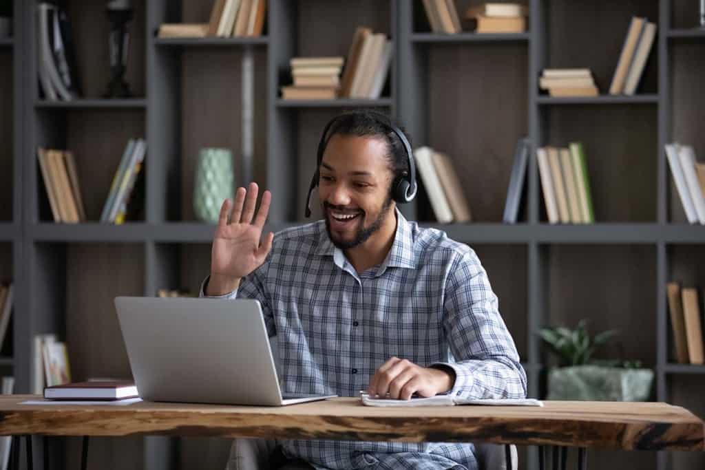 Un tip hipster african, prietenos și fericit, în căști cu microfon, salutând mâna la laptop, zâmbind, râzând, vorbind la o conferință video, folosind computerul pentru comunicare virtuală online de afaceri