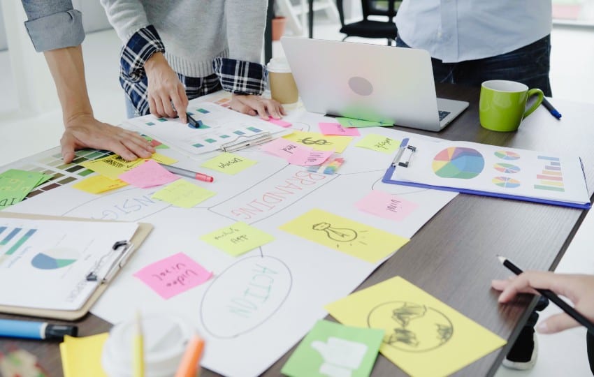 people brainstorming on a desk with paper, laptops, and clip boards around.