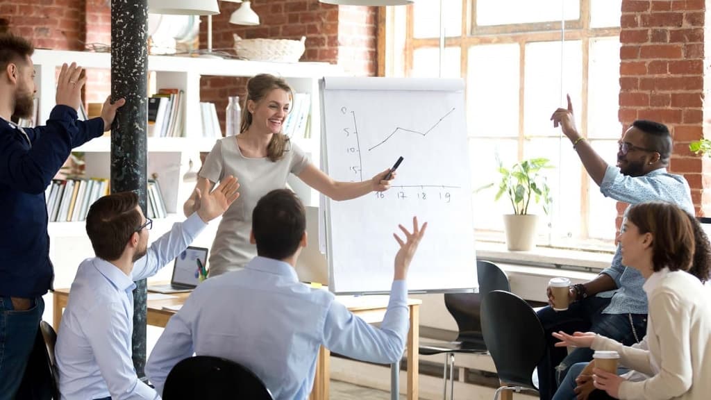 Successful cheerful businesswoman company head presenting new project to multi-ethnic young employees, diverse people voting raising hands or business coach giving presentation to clients in boardroom